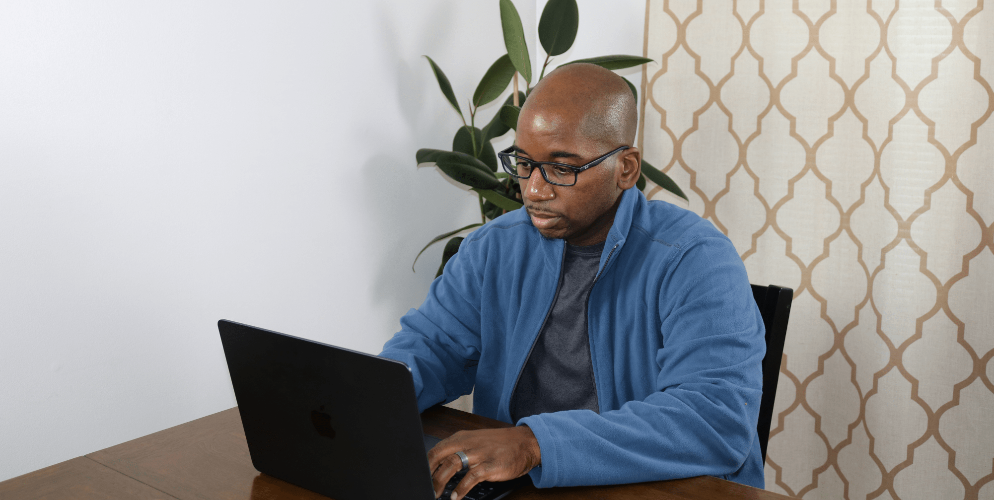 Rodney, a focused professional, works on his laptop at a wooden desk with a large potted plant and stylish backdrop.